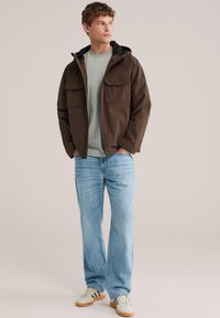 Jonge man die een bruine capuchonjas draagt, een lichtgrijs t-shirt, lichtblauwe spijkerbroek en witte sneakers met bruine strepen, staand met handen in de zakken.