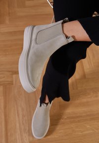 Helle graue Wildleder-Ankle-Boots mit elastischen Seitenpaneelen und einer Gummisohle, auf einem Holzboden platziert, getragen mit dunkler Kleidung.