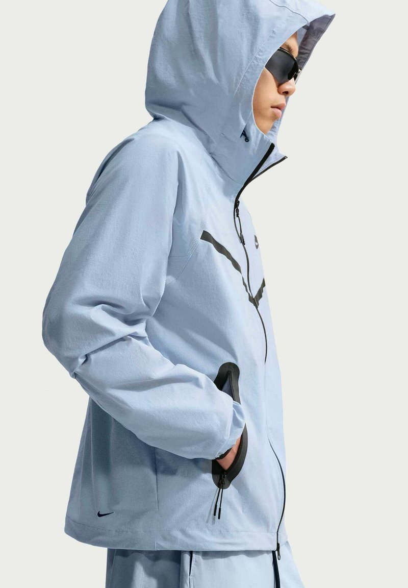 Person trägt hellblaue Kapuzenjacke und passende Hose mit Händen in den Reißverschlusstaschen, schaut nach rechts, trägt dunkle Sonnenbrille.