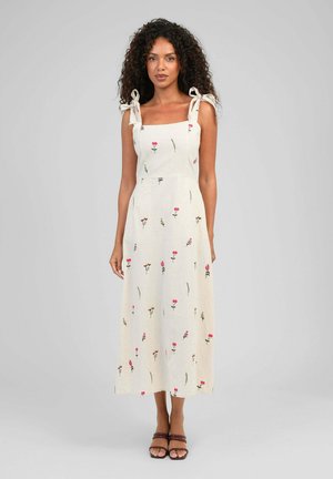 Femme aux cheveux bouclés portant une robe midi crème avec de fines bretelles et un petit motif floral, debout devant un fond gris.