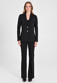 Completo su misura nero con giacca aderente dotata di due bottoni dorati e uno spacco, abbinato a pantaloni svasati. Tessuto di trama liscia.