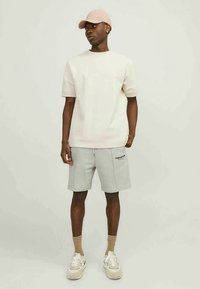 Uomo che indossa un berretto beige, una camicia a maniche corte bianco sporco, pantaloni corti grigi con il testo "Original Ride", calzini beige e sneakers bianche, in piedi contro uno sfondo semplice.