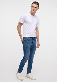Camiseta blanca de manga corta con cuello redondo, combinada con unos jeans de denim azul claro y zapatillas blancas. Diseño simple, corte casual, sin patrones.