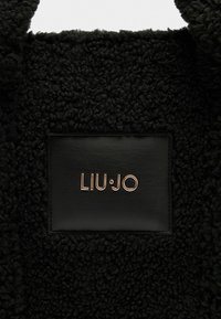 Un tissu noir texturé avec une apparence duveteuse présente un patch en cuir noir en relief avec "LIU·JO" en lettres dorées.