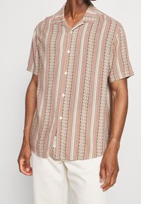 Chemise à manches courtes de couleur beige avec des rayures verticales rouges et crème. Dotée d'un col camp et de cinq boutons blancs sur le devant. Tissu texturé.