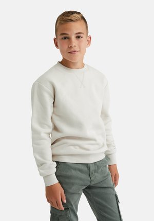 Sudadera beige clara de manga larga, con cuello redondo y puños acanalados. El modelo lleva pantalones cargo grises, que muestran un diseño relajado.
