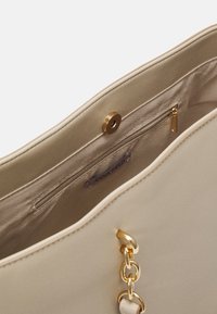 Intérieur de sac à main beige avec poche zippée, fermoir magnétique et détail de poignée chaîne dorée. L’étiquette indique « ANNA FIELD. »