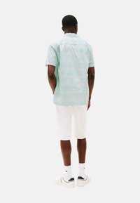 Camisa de mangas cortas de color verde menta con una textura de patrón sutil, combinada con pantalones cortos blancos y zapatillas, vista desde atrás.