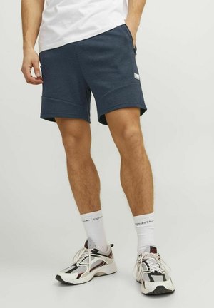 Pantalones cortos deportivos en azul marino, hechos de tela suave, con bolsillos laterales y un diseño deportivo. Se llevan con calcetines blancos y zapatillas deportivas de suela gruesa.