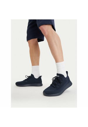 Hombre con pantalones cortos azul marino, calcetines blancos hasta la mitad de la pantorrilla y zapatillas deportivas negras, de pie sobre una superficie gris claro.
