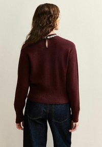 Maglione bordeaux con orlo a coste e maniche lunghe, caratterizzato da un dettaglio di apertura a chiave sul retro decorato con perline decorative. Indossato con denim scuro.