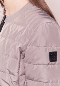 Vadderad jacka i ljus taupe, med rund halsringning, svart dragkedja och strukturerad finish. Inkluderar en svart logotyp patch på ärmen.