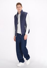 Mornarsko plavi quilted prsluk s visokim ovratnikom, zatvaranjem na zip, i bijelim detaljima, uparen s širokim plavim trapericama i bijelim tenisicama.