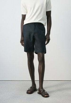 Uomo con camicia bianca a maniche corte, pantaloncini scuri fino al ginocchio e sandali di pelle marrone scuro in piedi contro un muro semplice.