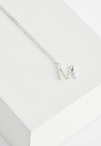 Ezüst nyaklánc, amelyen az "M" betű alakú függő látható, fehér háttér előtt. A lánc láncos kialakítású.