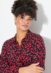 Schwarzes Button-up Hemd mit einem roten Herzmuster, langen Ärmeln und einem strukturierten Kragen. Glatte Textur mit dekorativen Hardware-Akzenten.