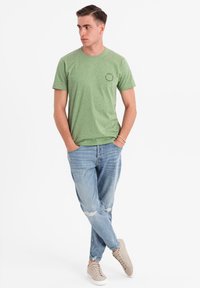 Camiseta verde de manga corta con un patrón jaspeado, cuello redondo y un pequeño logo. Combinada con jeans desgastados de color azul claro y zapatillas beige.