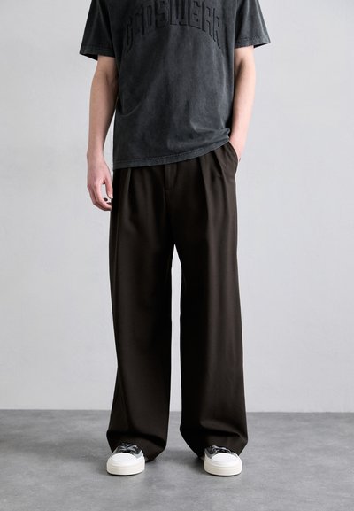T-shirt gris avec texte en relief et pantalon taille haute marron à coupe décontractée. Le modèle porte des baskets blanches avec des accents noirs.