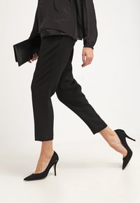 Svarta högklackade skor med spetstå, bärs med åtsittande svarta byxor och en lös svart top. Outfiten har en stilren, monokrom design.