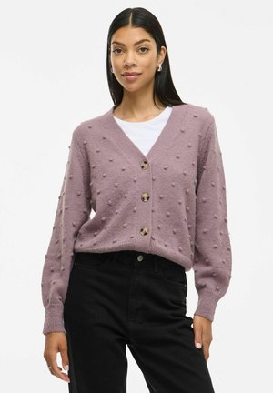 Cardigan lilla lavorato a maglia con texture gonfiata, scollatura a v e tre bottoni. Abbinato a una camicia bianca e pantaloni neri.