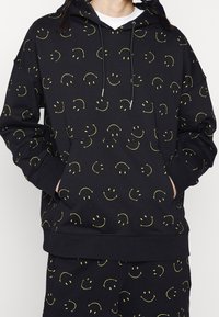 Black hoodie made of soft fabric, featuring an all-over pattern of yellow smiley faces, with a front pocket and drawstring hood.