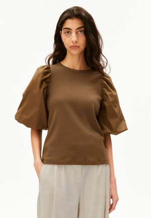 Vrouw met lang donker haar die een bruine top met pofmouwen en lichtbeige broek draagt, staand met één hand in de broekzak tegen een witte achtergrond.