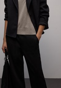 Veste noire cintrée portée sur un pull en maille beige, associée à un pantalon ample noir. Sac texturé noir avec des accents de ferrures argentées.