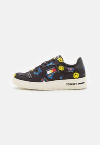 Zapatilla negra de Tommy Jeans con impresiones de cara sonriente y flores, suela blanca y la marca "TOMMY" en el lado y en la lengüeta.