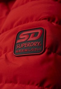Czerwona kurtka puchowa o pikowanej fakturze z czarną i czerwoną gumową naszywką z logo "SUPERDRY" oraz "SD" zaakcentowaną w rogu.