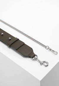 Cinturino in pelle verde oliva con hardware argentato, caratterizzato da borchie, attaccato a un guinzaglio in catena metallica. Entrambi i lati hanno ganci a artiglio e a moschettone.