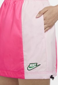 Shorts Nike avec un design divisé en rose vif et rose doux, ornés d'un logo vert brodé en bas à gauche. Matériau léger.