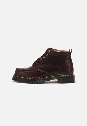 Σκούρο καφέ δερμάτινο ankle boot με δέσιμο μπροστά, κίτρινο ράψιμο γύρω από τη σόλα και λουράκι στο πίσω μέρος.
