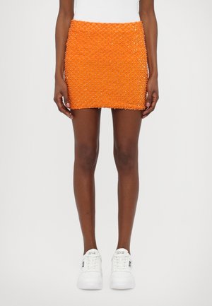 Mini-jupe orange avec surface texturée ornée de perles rondes transparentes ; associée à des baskets blanches, présentant un look décontracté.