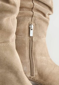 Beige suède laarzen tot de knie hebben een zijrits en geplooid stofdetail, met zichtbare stiksels langs de naden voor extra textuur.