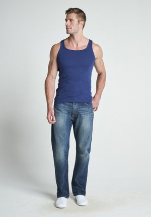 Man in blauw tanktop, vervaagde blauwe spijkerbroek en witte schoenen staat zijwaarts kijkend tegen een effen witte achtergrond.