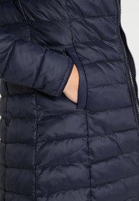 Tmavě modrý vatovaný kabát s prošívaným designem, bočními kapsami a zapínáním na zip. Materiál vypadá hladce a voděodolně.