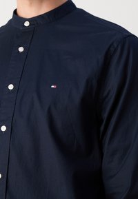 Homme portant une chemise bleu marine foncé avec un col officier, des boutons blancs sur le devant et un petit logo rouge, blanc et bleu sur la poitrine.