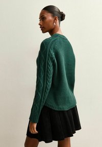 Zielony sweter z dzianiny z detalami w warkocz na jednym rękawie, w zestawie z czarną spódnicą z sztruksu. Miękka faktura, okrągły dekolt, luźny krój.