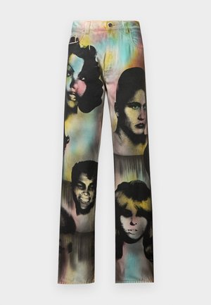 Mann trägt ein gelbes, marineblaues, braunes und weiß gestreiftes Poloshirt, mehrfarbige Jeans mit großen schwarz-weißen Gesichtsaufdrucken und schwarze Schuhe.