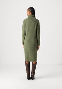 Πράσινο πλεκτό φόρεμα με ραβδώσεις, ψηλό γιακά, μακριά μανίκια και εφαρμοστή σιλουέτα, συνδυασμένο με καφέ δερμάτινες μπότες μέχρι το γόνατο.