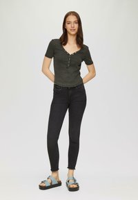 Donkergrijze, aansluitende top met korte mouwen en een V-hals, voorzien van knoopdetails. Gecombineerd met zwarte skinny jeans en blauwe plateausandalen. Minimalistisch ontwerp.