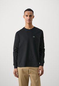 Black long-sleeve t-shirt made of cotton, featuring a small green Lacoste logo on the left chest. Paired with khaki pants.