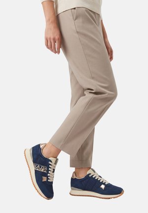 Persoon draagt beige broek en donkerblauwe sneakers met lichtbruine zolen, staand met één voet licht opgetild.