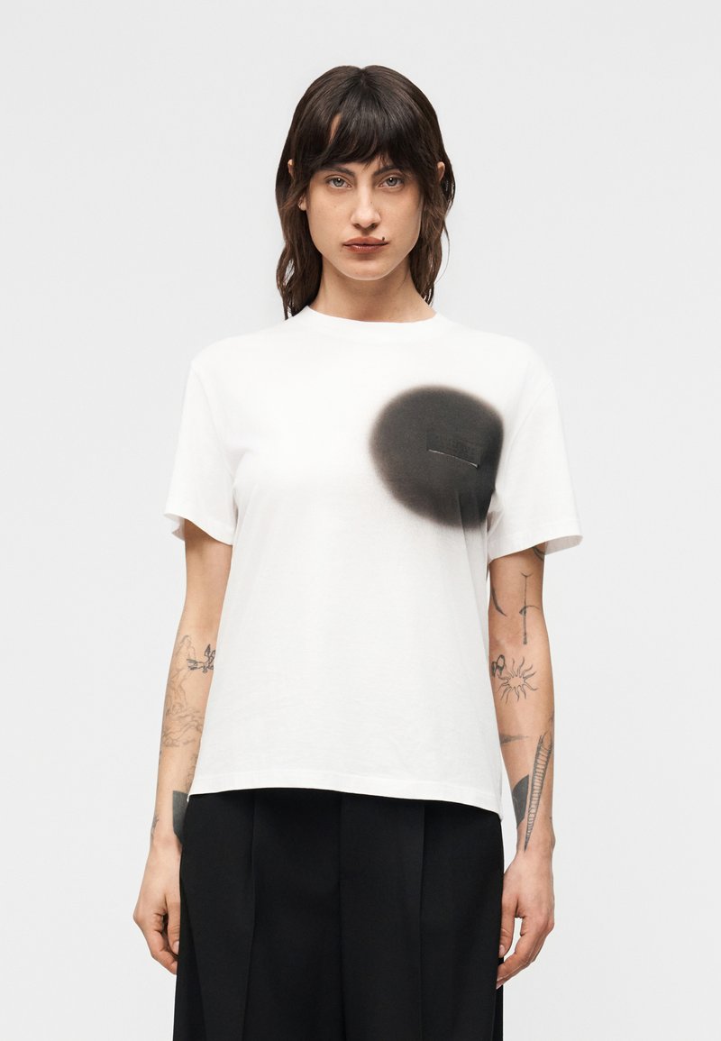 Γυναίκα με σκούρα μαλλιά που φοράε ι λευκό μπλουζάκι με μαύρο κύκλο σπρέι κοντά στο στήθος, μαύρο παντελόνι και εμφανή τατουάζ στα χέρια, στέκεται στραμμένη προς τα εμπρός.