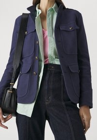 Chaqueta azul marino con tela texturizada, cierre de botones y cuatro bolsillos frontales, superpuesta a una camisa verde menta clara y pantalones de mezclilla negros.