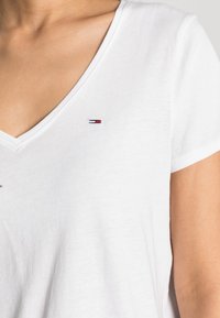 Camiseta de algodón blanca con cuello en V que presenta un pequeño logotipo en rojo, blanco y azul en el lado izquierdo del pecho. Textura suave con mangas cortas.