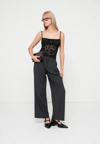 Schwarzes Blumen-Spitzenoberteil mit transparenter Struktur, hoch taillierte graue Hose und schwarze spitz zulaufende High Heels. Hält eine kleine, dunkle Clutch.