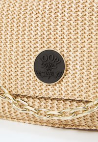 Beżowa torba tekstylna z plecionki z czarną naszywką logo, z złotym pasekiem łańcuchowym i teksturowanym, skomplikowanym wzorem na powierzchni.