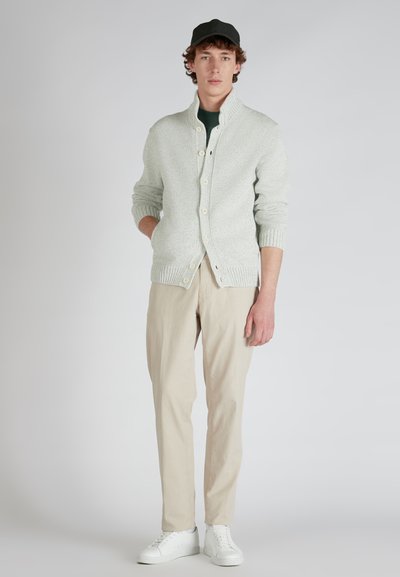  lichtgroene gebreide cardigan met hoge kraag, knoopsluiting en geribde mouwen, gecombineerd met beige broeken en witte sneakers.