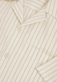 Cămașă de pijama stripedă albă cu dungi bej înguste. Are un guler, două butoane și o buzunar pe piept. Material moale, texturat.